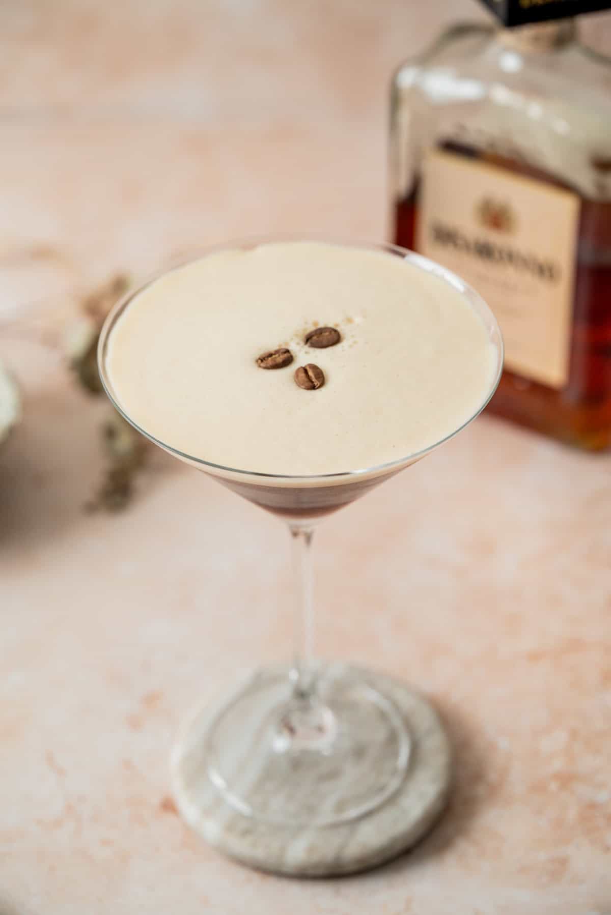 A creamy Amaretto Espresso Martini garnished with three coffee beans in a martini glass on a marble coaster, with a blurred bottle of liqueur in the background.
