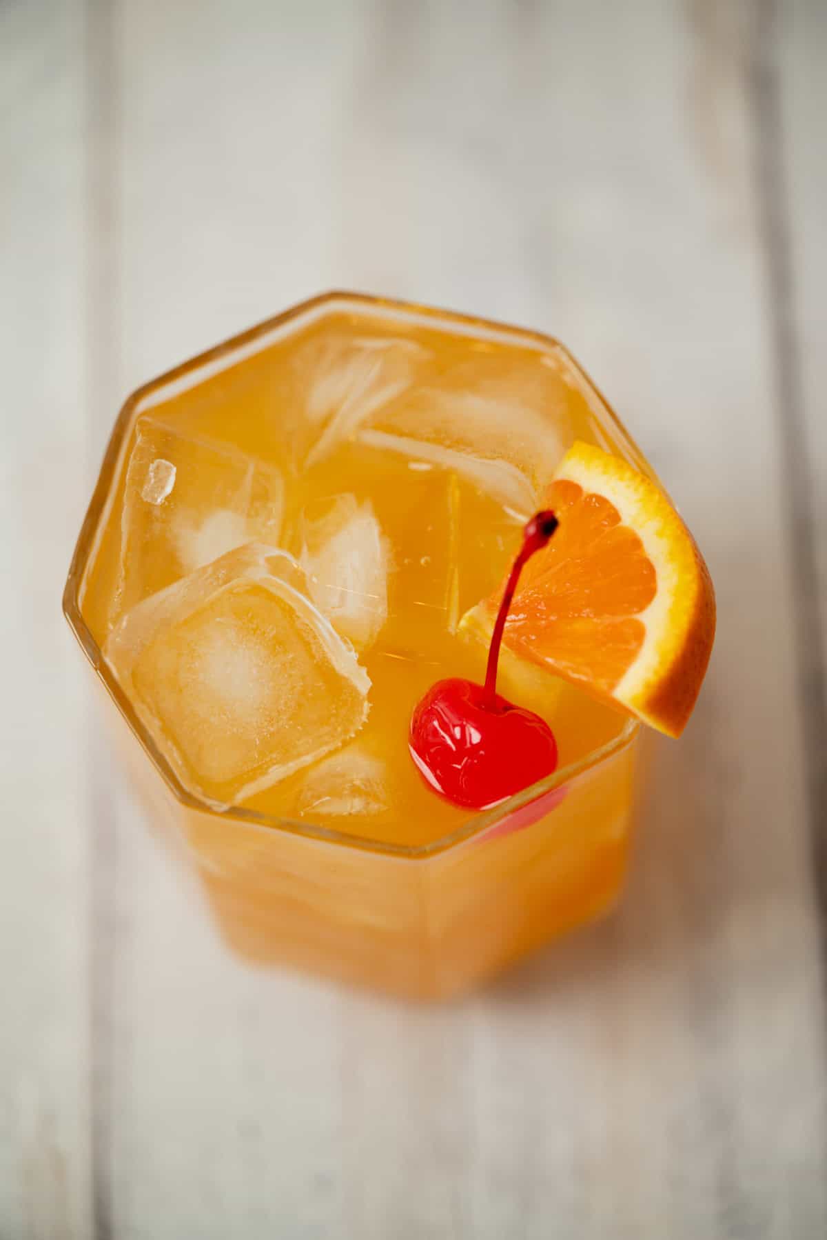 A glass of amaretto stone sour with ice cubes, garnished with a maraschino cherry and an orange slice, sits on a light wooden surface.