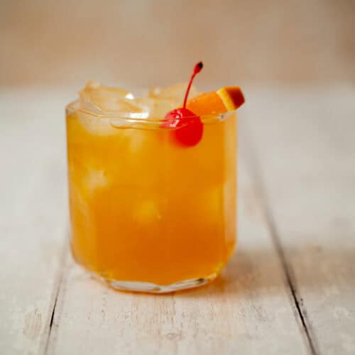 A glass of amaretto stone sour with ice, garnished with a red cherry and an orange slice, sits on a light wooden surface.