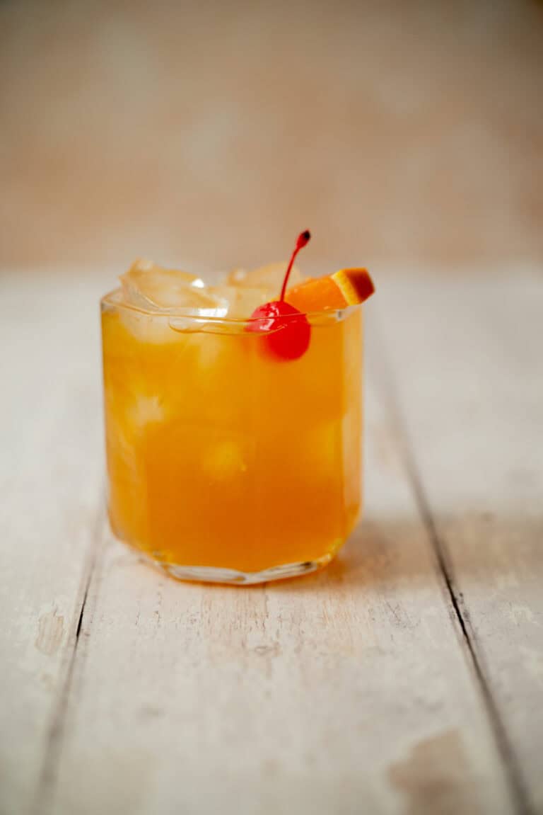 A glass of amaretto stone sour with ice, garnished with a red cherry and an orange slice, sits on a light wooden surface.