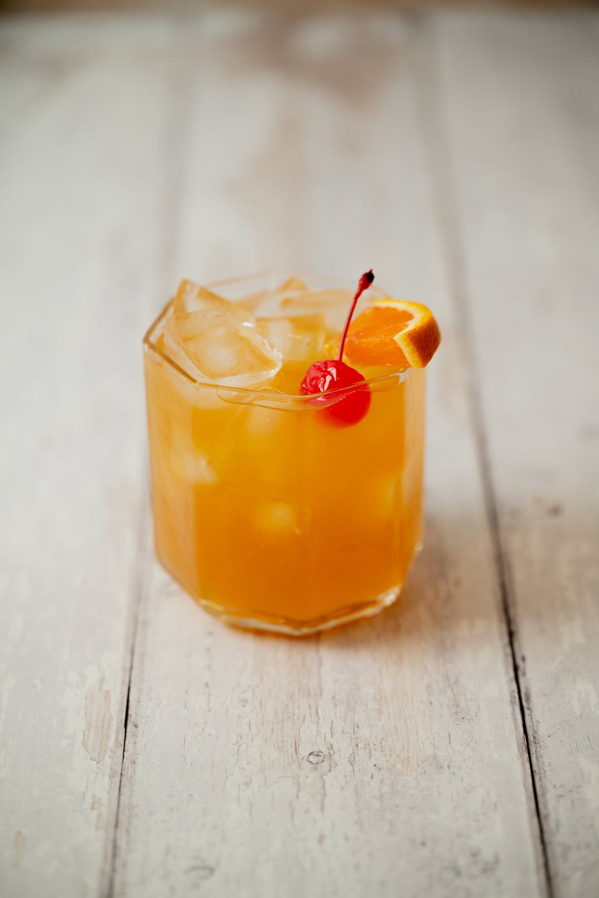 A glass of orange-colored amaretto stone sour with ice cubes, garnished with a red cherry and an orange slice, sits on a rustic white wooden surface.