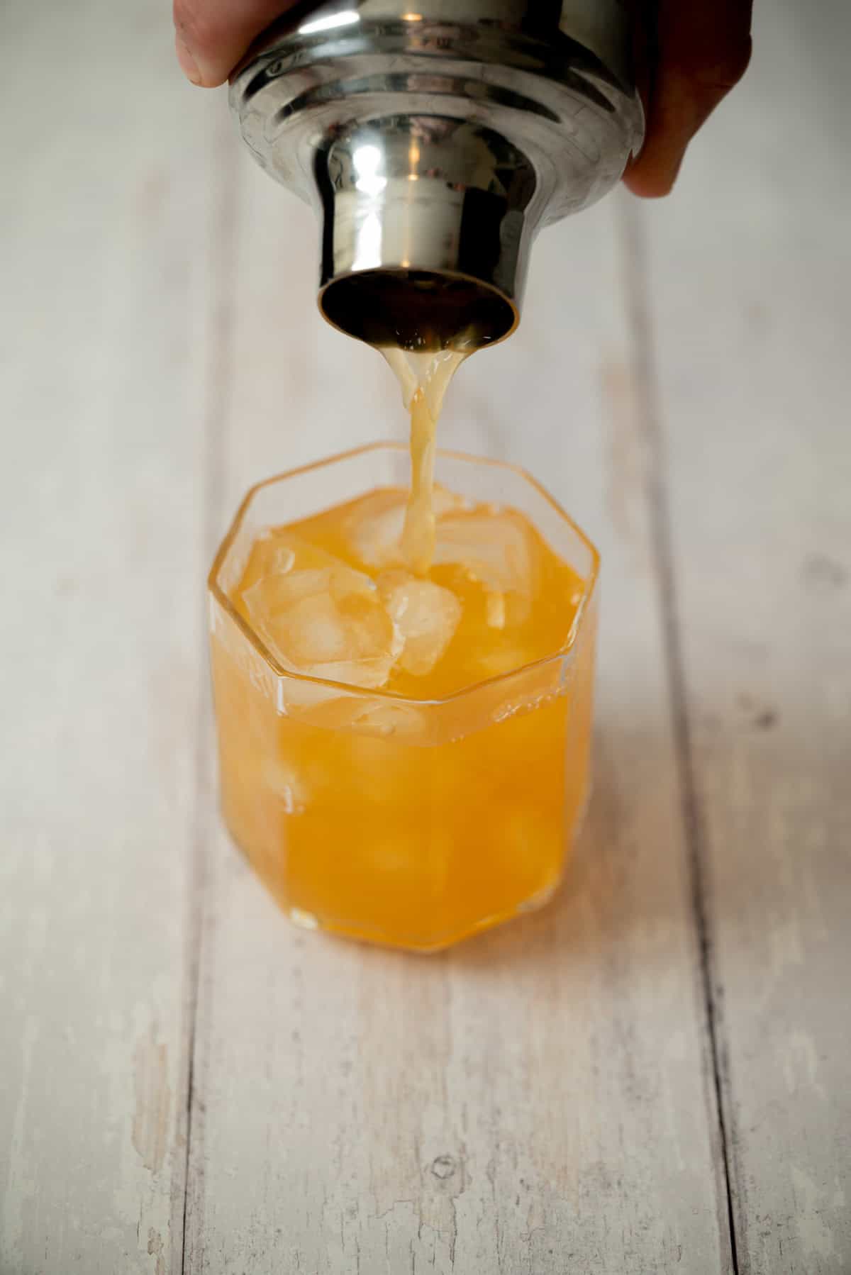 A close-up of a hand pouring an amaretto stone sour from a metal shaker into a glass filled with ice, set on a light wooden surface.