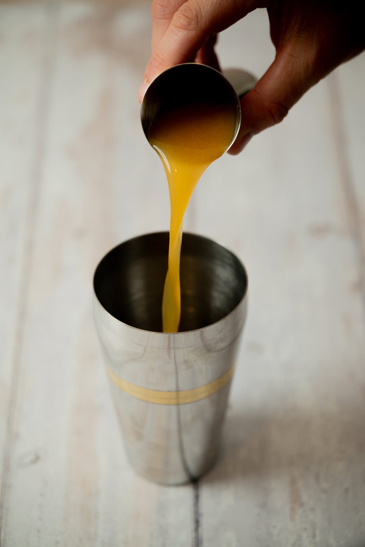 A hand pours orange liquid for an amaretto stone sour from a small metal jigger into a stainless steel cocktail shaker on a light wooden surface.