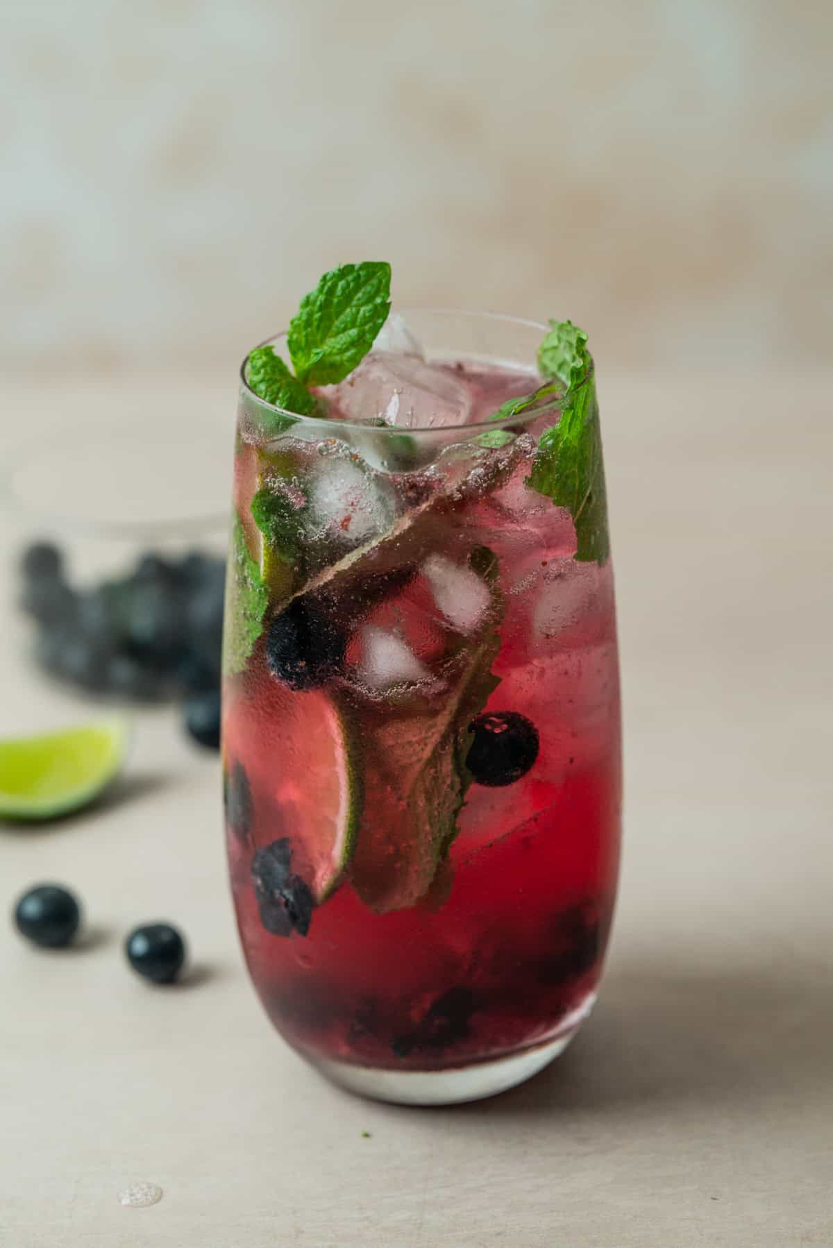A tall glass filled with a vibrant pinkish-red blueberry mojito mocktail, ice cubes, blueberries, lime slices, and fresh mint leaves, with extra blueberries, a lime wedge, and a glass bowl blurred in the background.