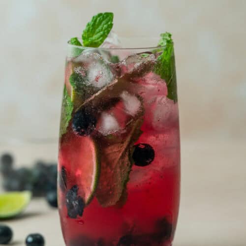 A tall glass filled with a vibrant Blueberry Mojito Mocktail, brimming with ice cubes, fresh mint leaves, lime slices, and whole blueberries, set on a light surface with scattered blueberries and lime wedges nearby.