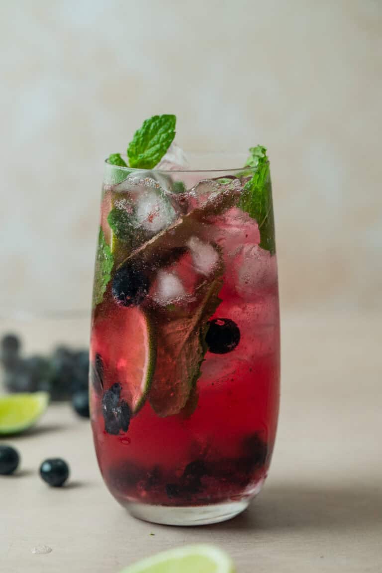 A tall glass filled with a vibrant Blueberry Mojito Mocktail, brimming with ice cubes, fresh mint leaves, lime slices, and whole blueberries, set on a light surface with scattered blueberries and lime wedges nearby.