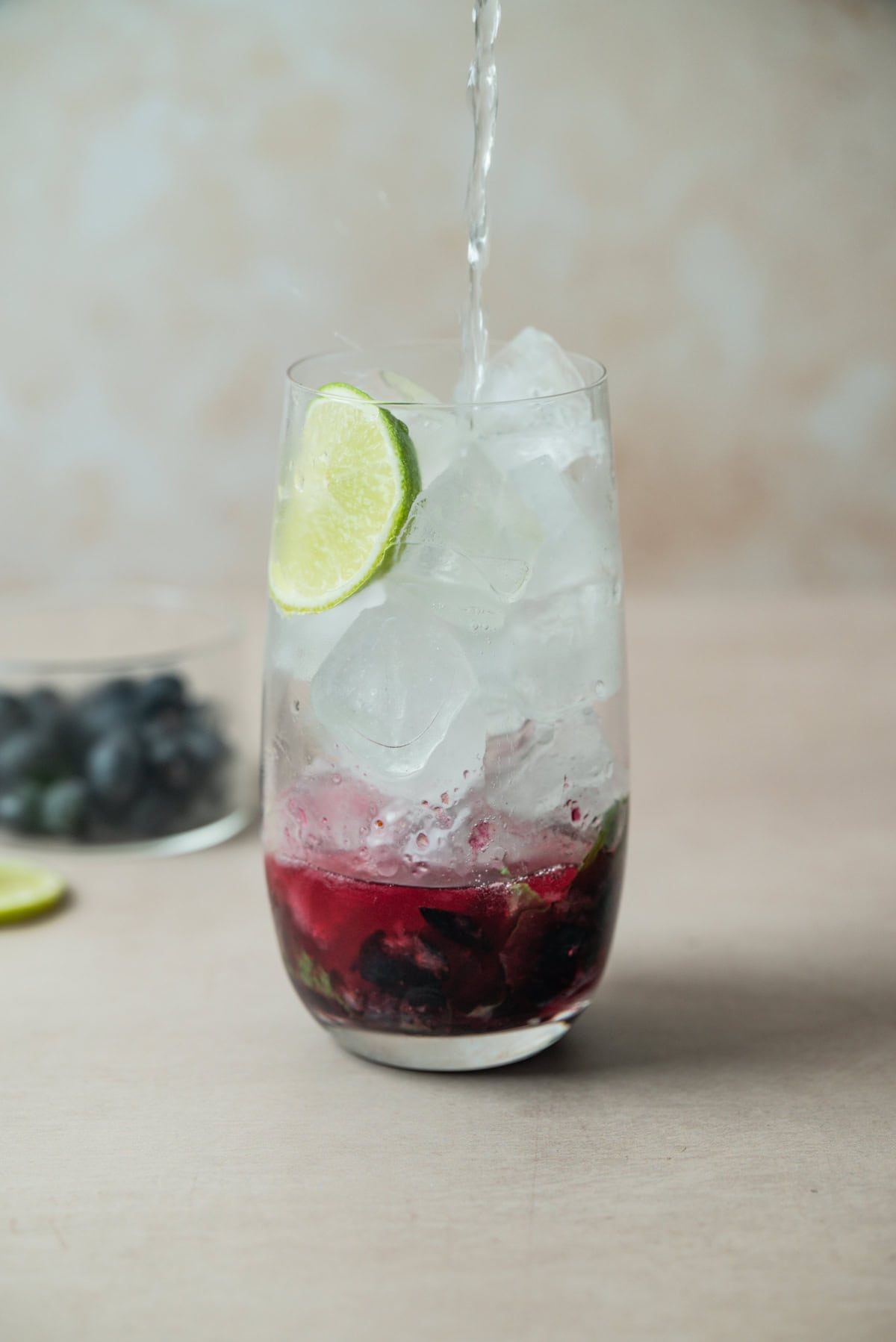 A glass filled with ice, sliced lime, and a red fruit mixture at the bottom is being topped with a clear liquid—perfect for a refreshing Blueberry mojito Mocktail. In the background, dark grapes and a slice of lime rest on a light surface.