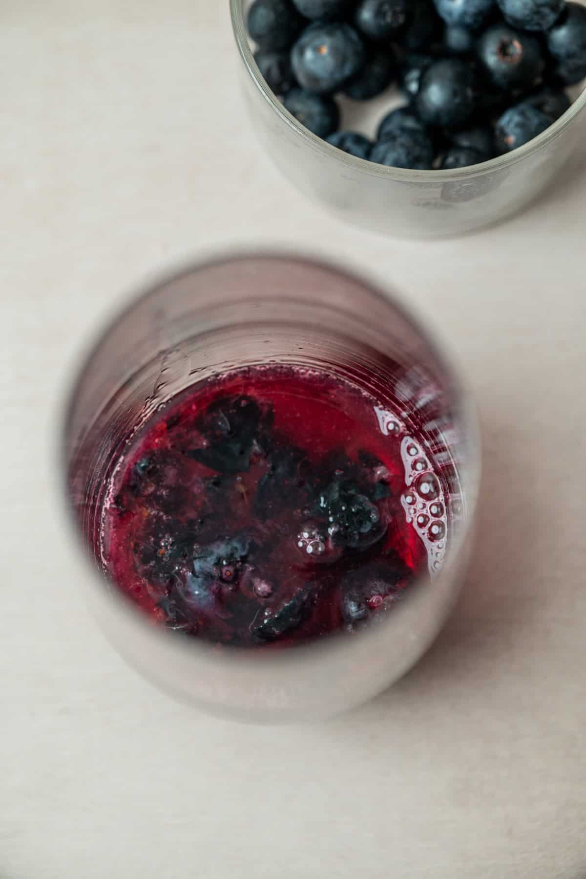 A close-up view of a glass containing a dark red blueberry mojito mocktail, with whole blueberries visible. Behind it, there is a second glass bowl filled with fresh blueberries on a light surface.