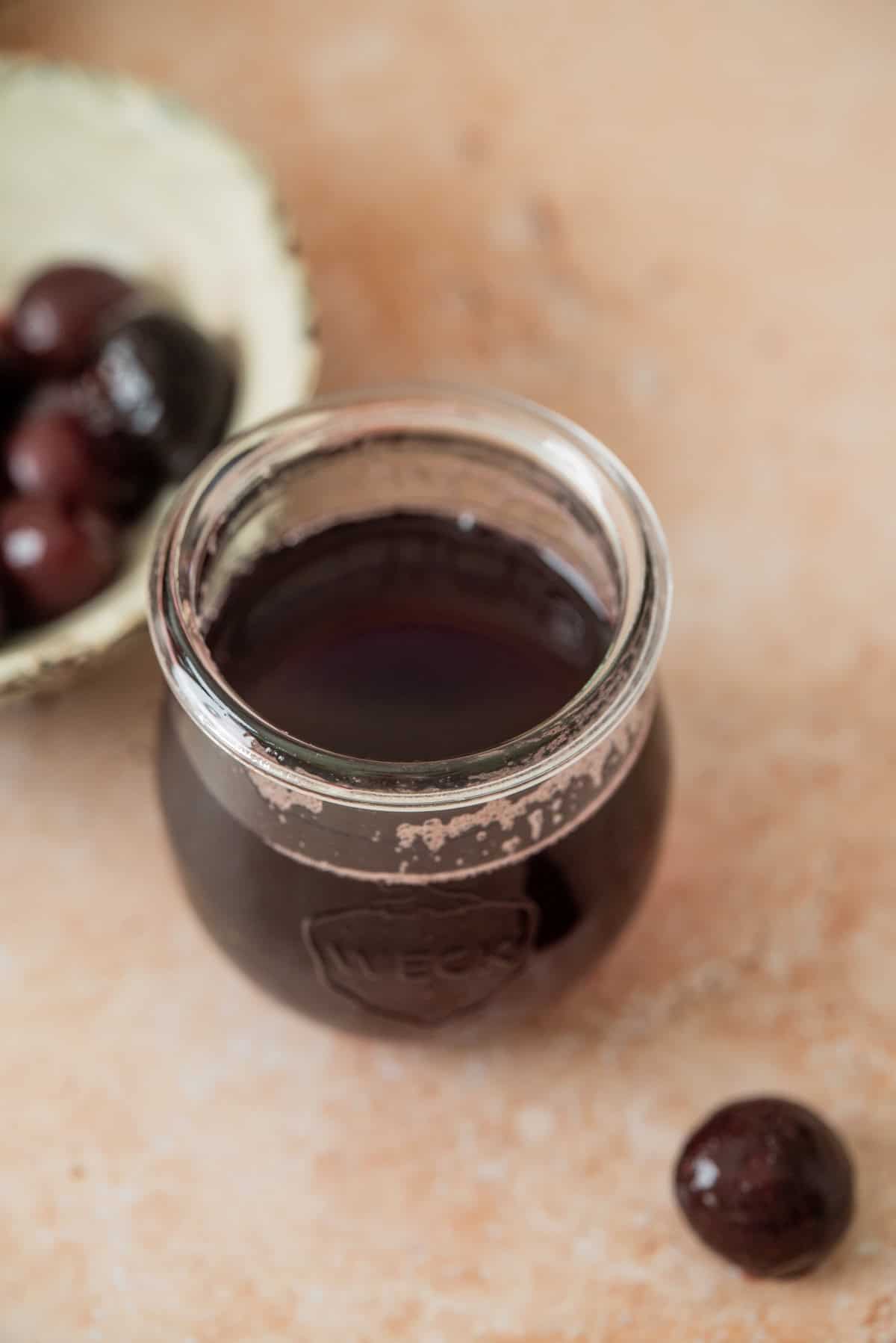 A glass jar filled with dark cherry simple syrup sits on a light surface. Beside it is a whole cherry and a bowl containing several cherries.