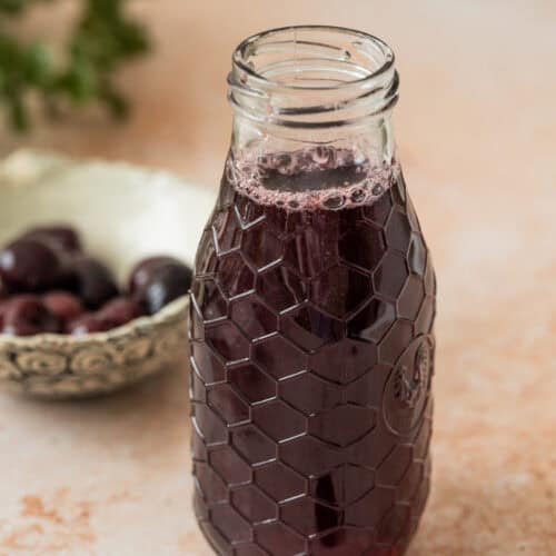 A glass bottle with a honeycomb pattern holds dark purple cherry simple syrup. In the background, a bowl of cherries and a few loose cherries rest on a light-colored surface.