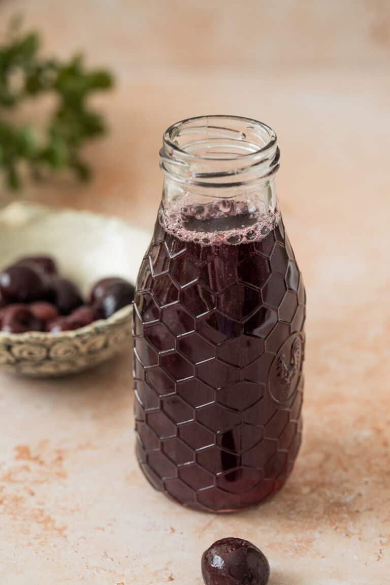A glass bottle with a honeycomb pattern holds dark purple cherry simple syrup. In the background, a bowl of cherries and a few loose cherries rest on a light-colored surface.