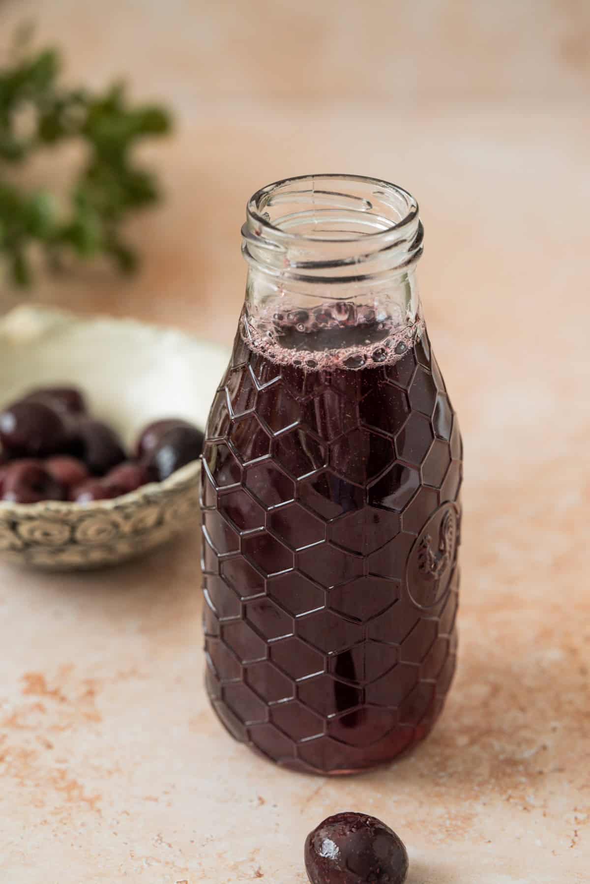 A glass bottle with a honeycomb pattern holds dark purple cherry simple syrup. In the background, a bowl of cherries and a few loose cherries rest on a light-colored surface.