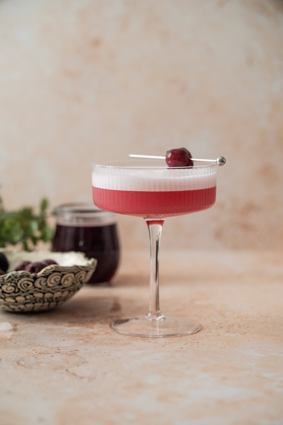 A pink cherry gin sour with a frothy foam top is served in a coupe glass, garnished with a dark cherry on a pick. In the background are a decorative bowl of cherries and a jar of dark liquid on a light stone surface.