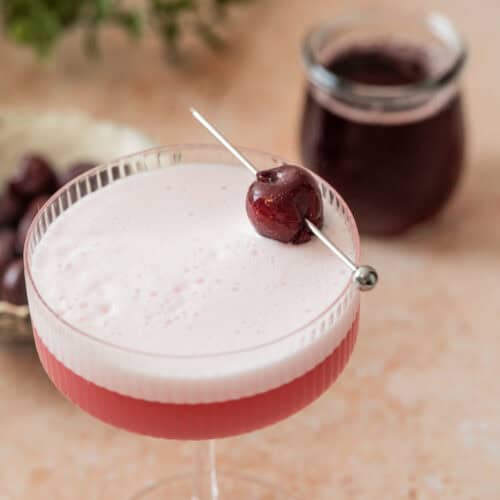 A frothy pink cherry gin sour sits in a coupe glass, garnished with a dark cherry on a cocktail pick. In the background, there’s a jar of dark liquid and a small dish filled with more cherries on the light tan surface.