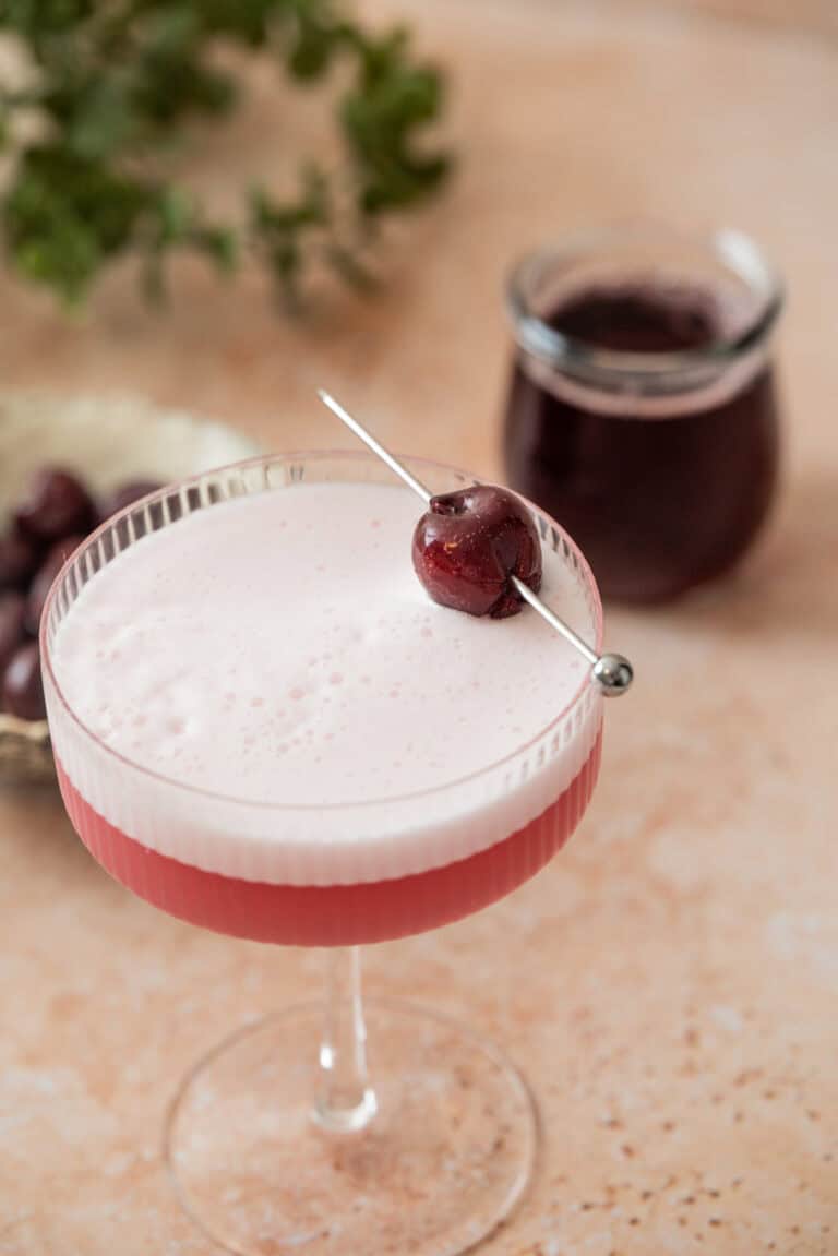 A frothy pink cherry gin sour sits in a coupe glass, garnished with a dark cherry on a cocktail pick. In the background, there’s a jar of dark liquid and a small dish filled with more cherries on the light tan surface.