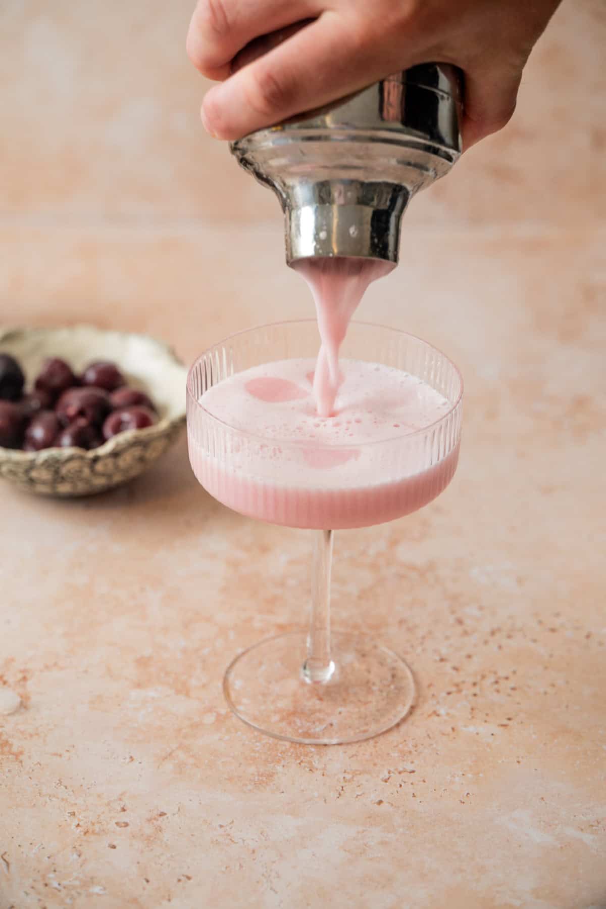 A hand pours a frothy pink cherry gin sour from a shaker into a glass. In the background, a small bowl of cherries rests on a light-colored countertop.