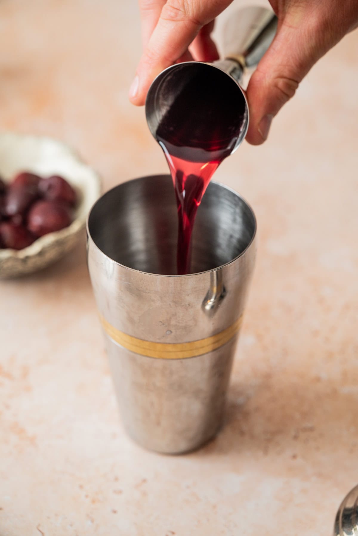 A hand pours red liquid for a cherry gin sour from a small measuring cup into a metal cocktail shaker; cherries in a bowl sit blurred in the background.