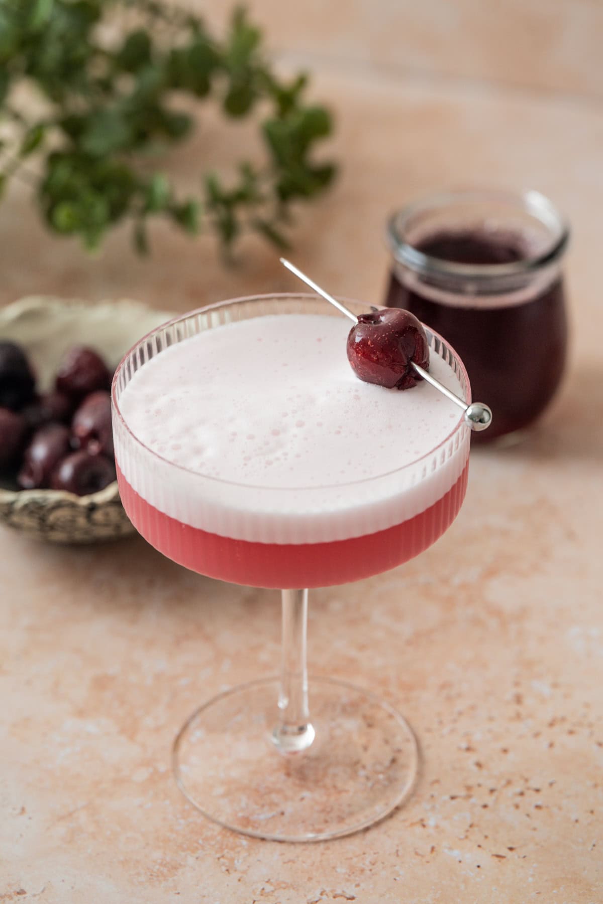A gorgeous pink cherry gin sour cocktail in a coupe glass, topped with a cherry on a pick. In the background, there’s a bowl of cherries, a jar of dark red liquid, and green leaves on a light beige surface.