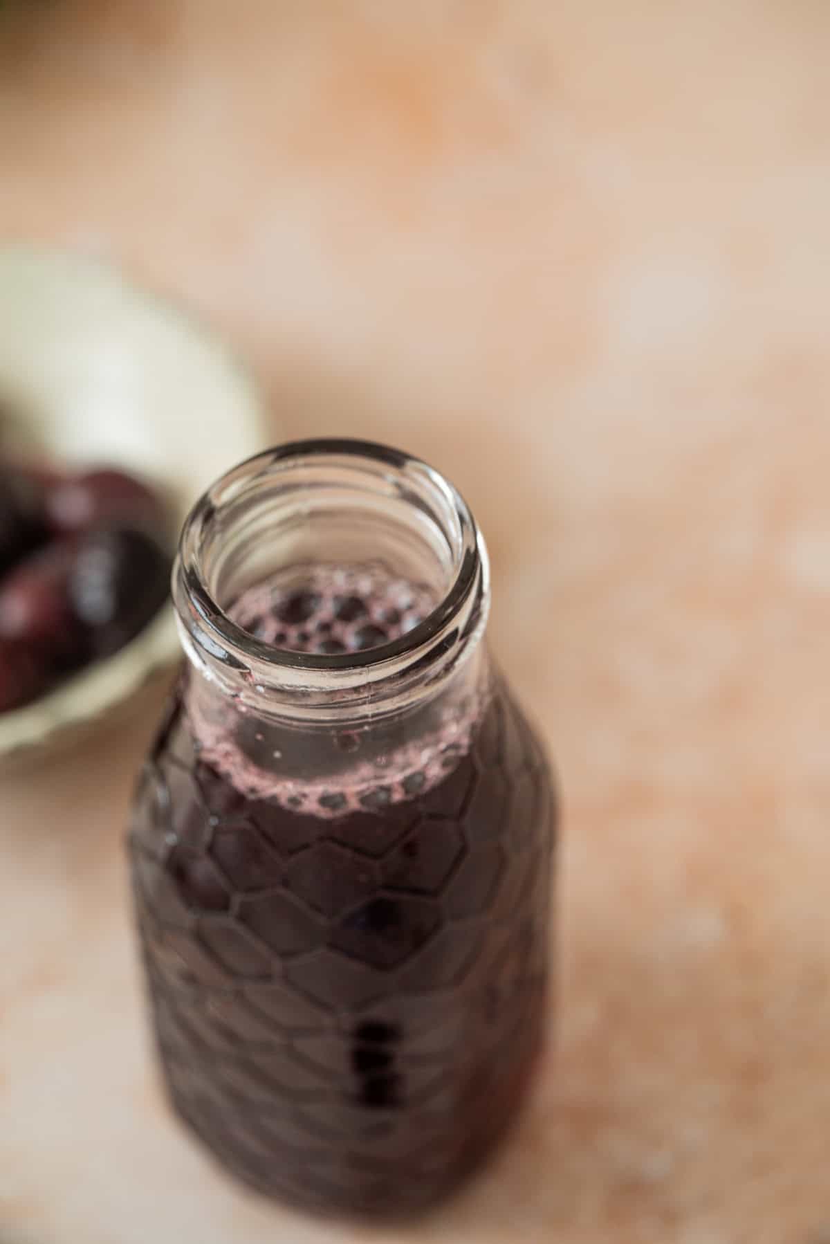A close-up of a glass bottle filled with dark purple cherry simple syrup, with a bowl of dark fruits blurred in the background on a light-colored surface.
