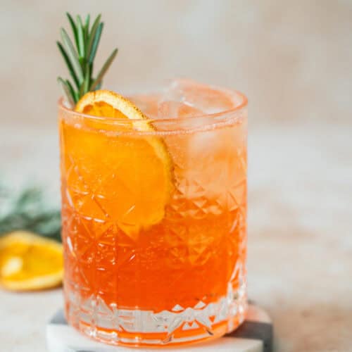 A sparkling Aperol Soda orange cocktail with ice, garnished with a sprig of rosemary and a dried orange slice, served in a patterned glass on a round marble coaster.