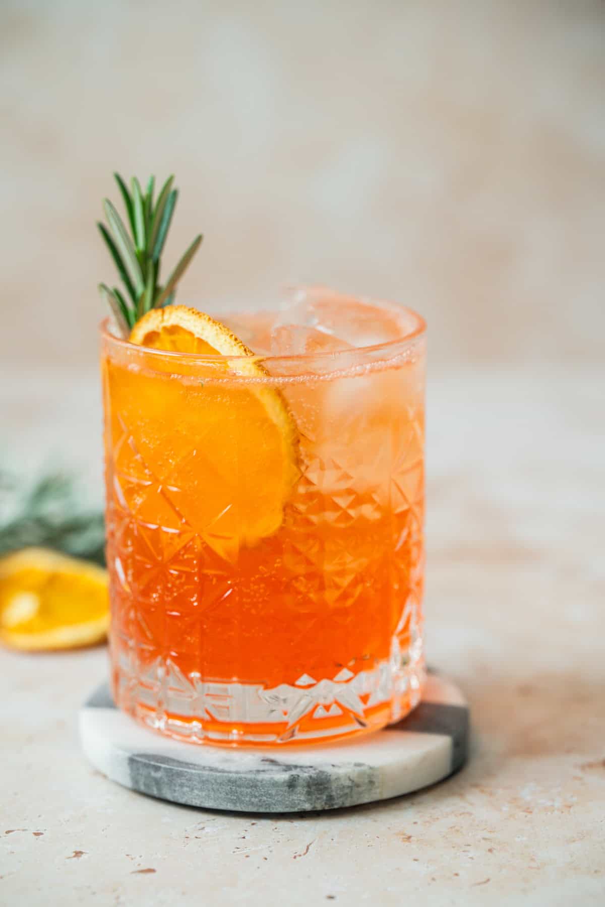 A sparkling Aperol Soda orange cocktail with ice, garnished with a sprig of rosemary and a dried orange slice, served in a patterned glass on a round marble coaster.