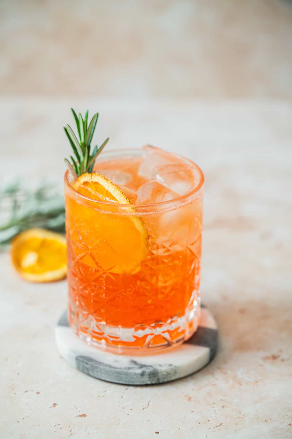 A glass of Aperol Soda cocktail with ice, garnished with an orange slice and a sprig of rosemary, sits on a round marble coaster against a light, blurred background.