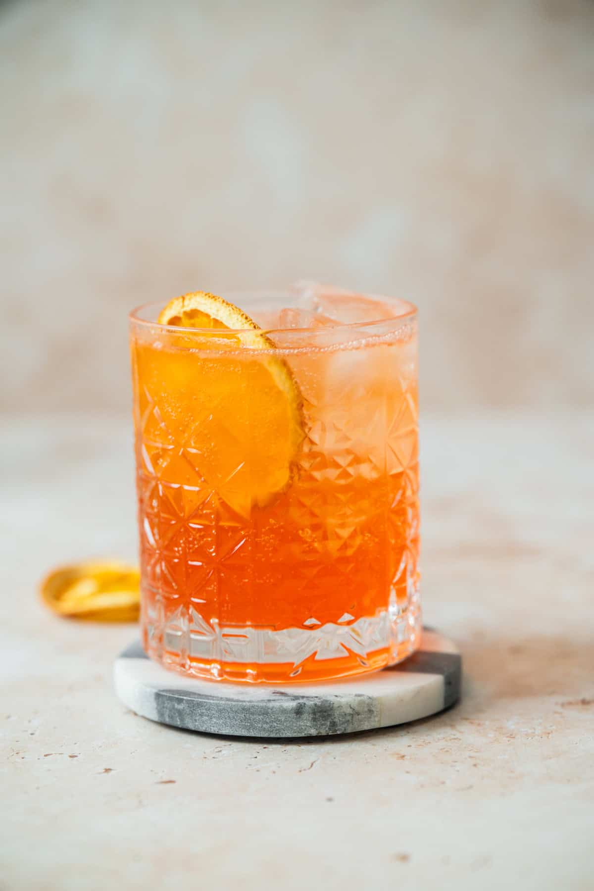 A glass filled with an Aperol Soda cocktail on ice, garnished with a dried orange slice, sits on a round marble coaster against a neutral background.