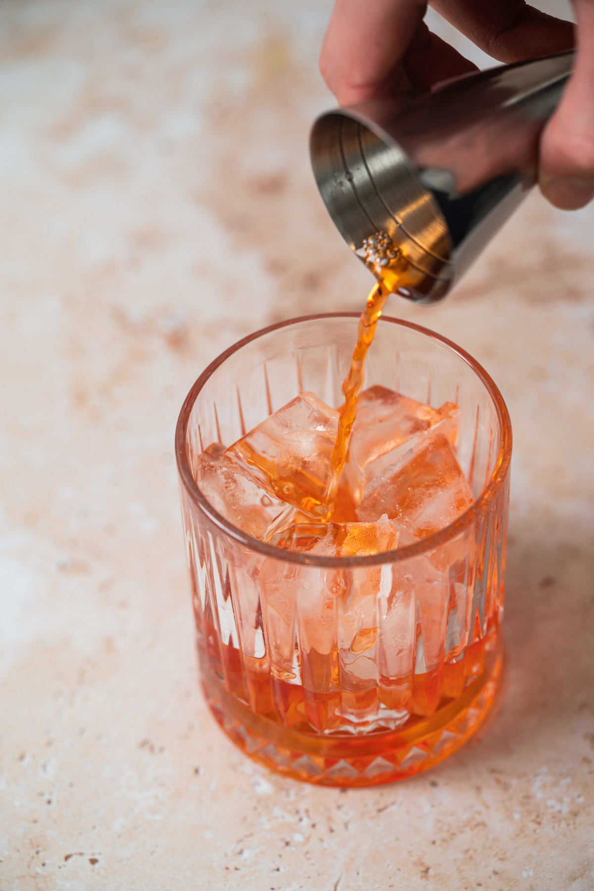 A hand pours an orange-colored Aperol Ginger Beer drink from a metal jigger into a glass filled with ice cubes, set on a light-colored surface.