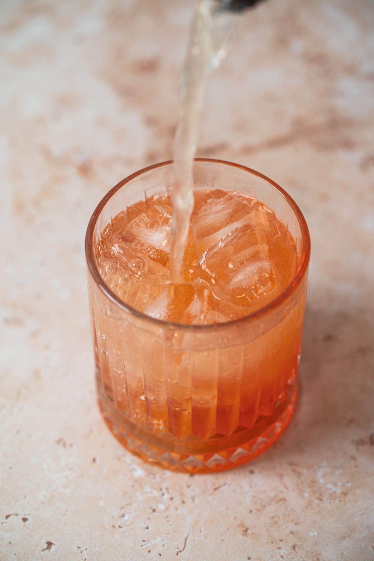 A clear, textured glass filled with ice cubes and an orange-colored Aperol Ginger Beer drink sits on a light, marbled surface as a liquid is being poured into it.