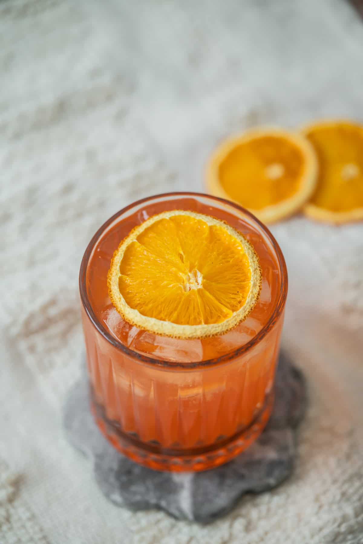 A glass of orange-colored Aperol Ginger Beer with ice, garnished with a round orange slice, sits on a dark coaster. Two more orange slices are in the blurred background on a light textured surface.