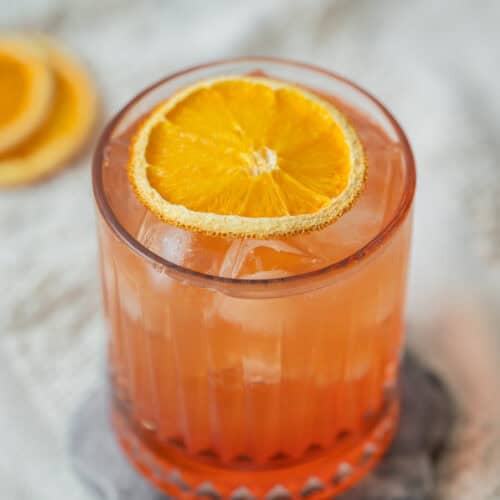 A glass of Aperol Ginger Beer cocktail with ice, garnished with a dried orange slice, sits on a stone coaster. Blurred orange slices are in the background on a textured fabric surface.