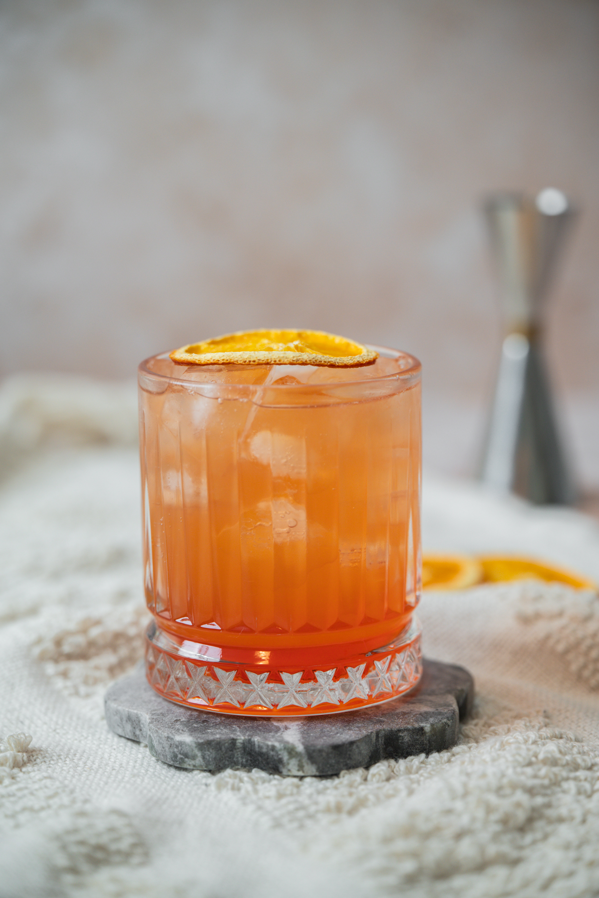A glass of orange-colored Aperol Ginger Beer cocktail with ice and an orange slice garnish sits on a stone coaster, with a blurred jigger and more orange slices in the background.
