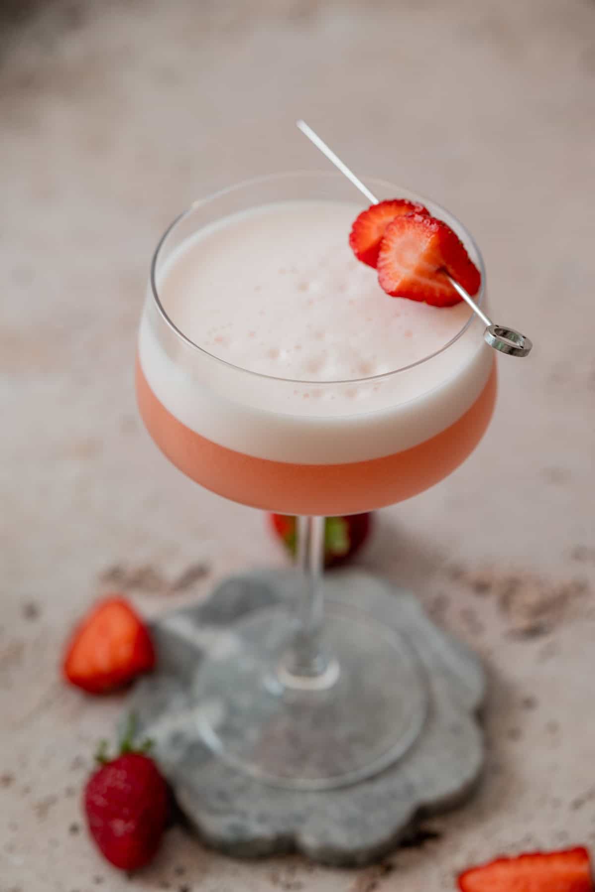 A pink, frothy Strawberry Gin Sour in a coupe glass, garnished with sliced strawberry on a cocktail pick. Fresh strawberries are placed beside the glass on a gray stone coaster.