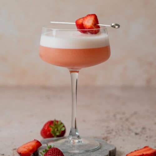 A pink Strawberry Gin Sour with a frothy top is served in a coupe glass, garnished with strawberry slices on a cocktail pick. Fresh strawberries are placed around the glass on a stone coaster and a light-colored surface.