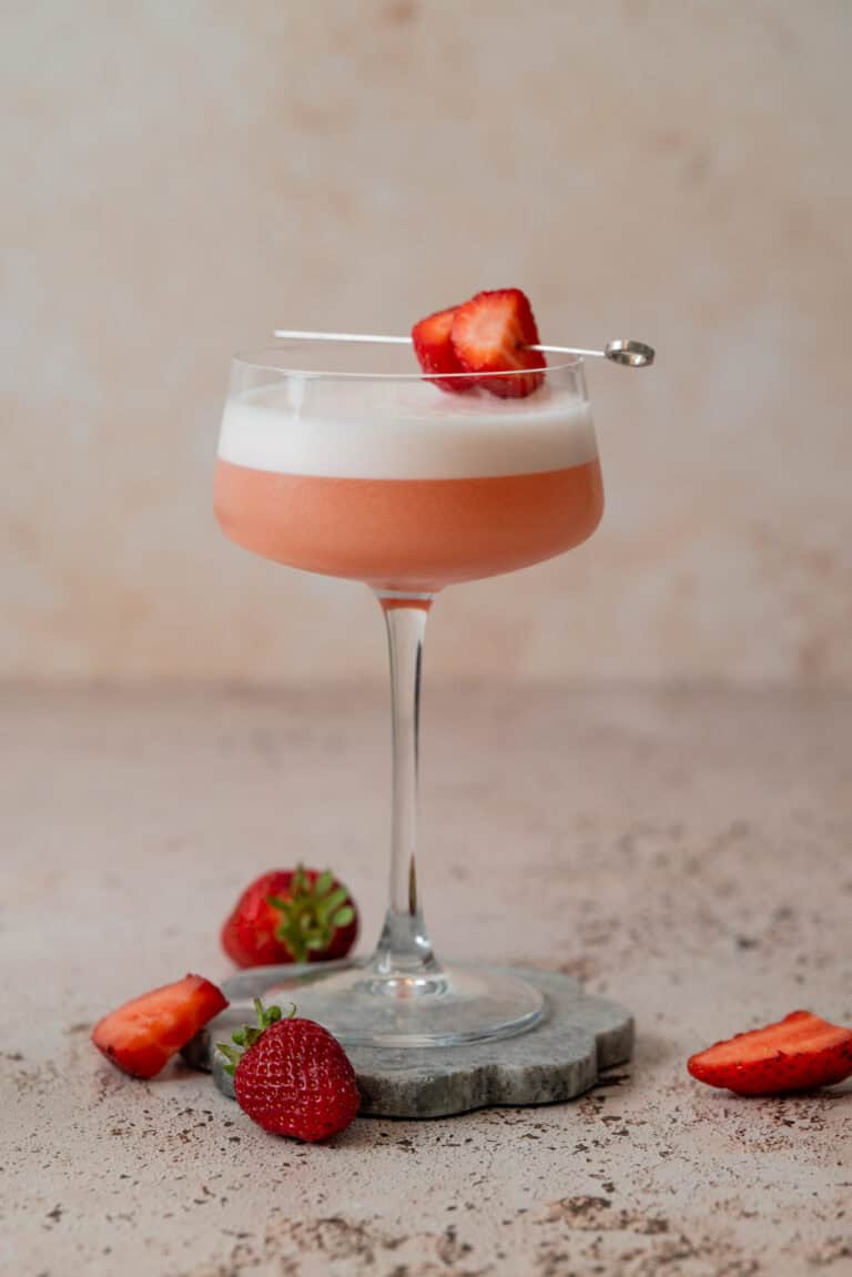 A pink Strawberry Gin Sour with a frothy top is served in a coupe glass, garnished with strawberry slices on a cocktail pick. Fresh strawberries are placed around the glass on a stone coaster and a light-colored surface.