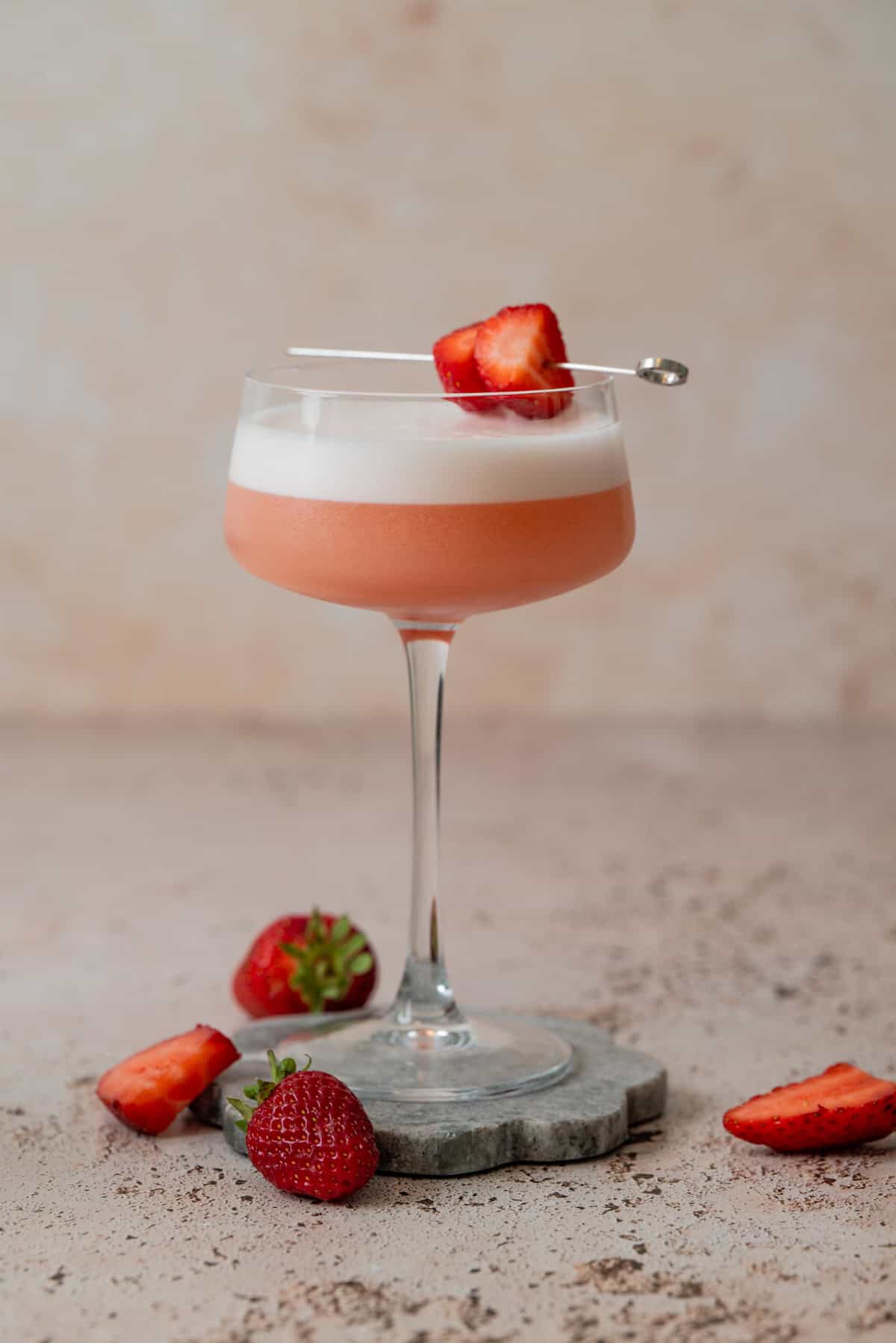 A pink Strawberry Gin Sour with a frothy top is served in a coupe glass, garnished with strawberry slices on a cocktail pick. Fresh strawberries are placed around the glass on a stone coaster and a light-colored surface.
