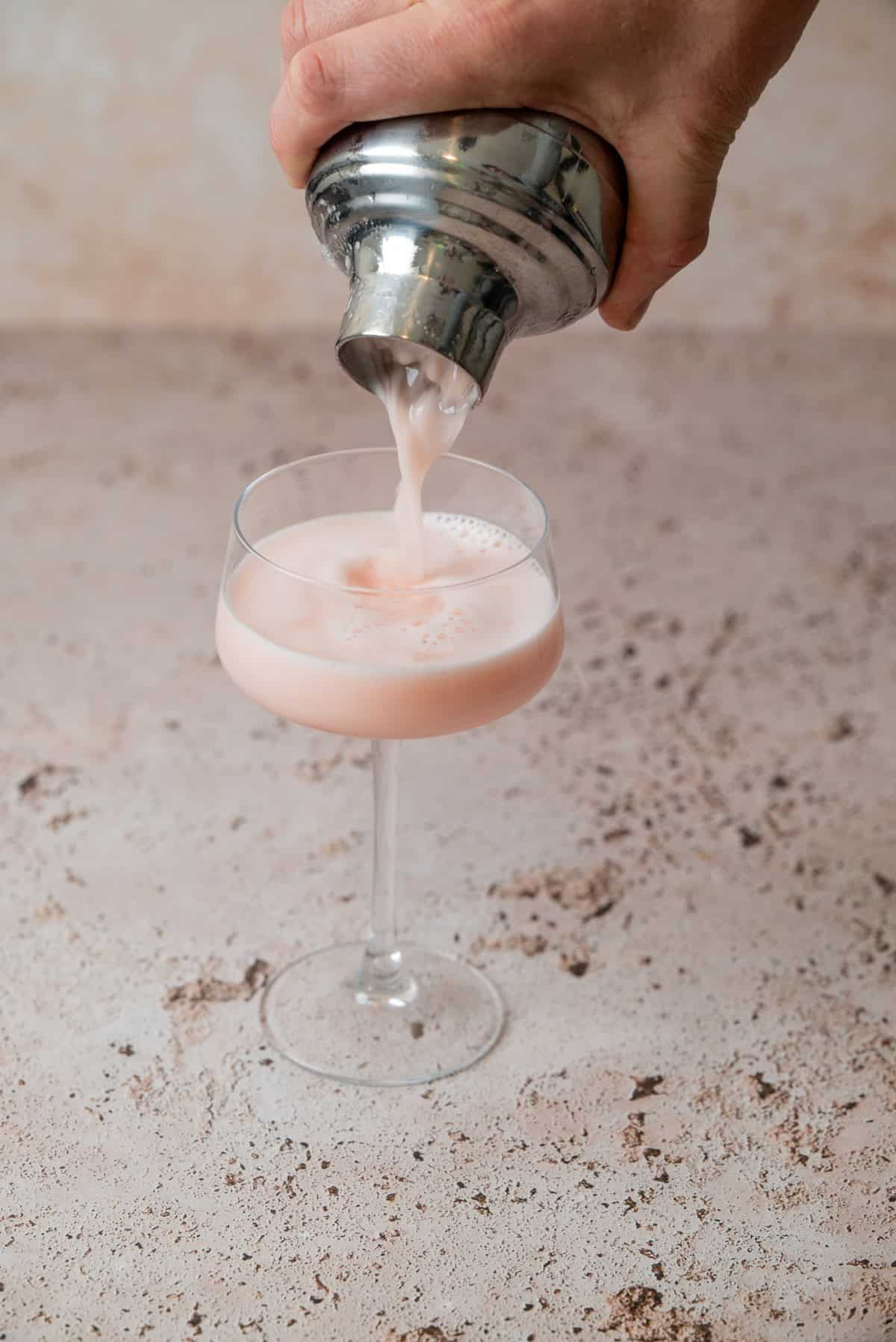 A hand pours a creamy pink Strawberry Gin Sour from a metal shaker into a stemmed glass on a speckled, light-colored surface.