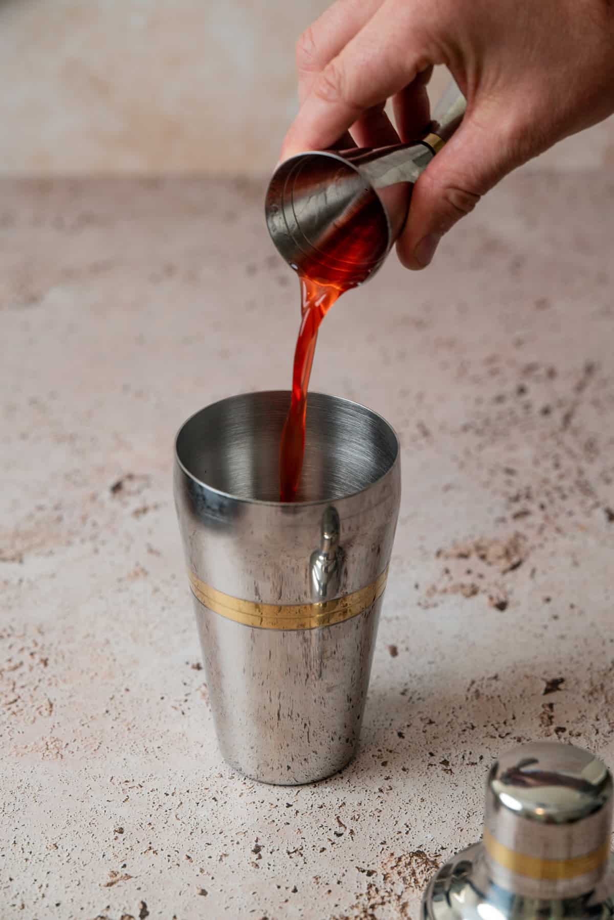 A hand pours a red liquid for a Strawberry Gin Sour from a metal jigger into a stainless steel cocktail shaker on a light textured surface, with the shaker’s lid resting nearby.