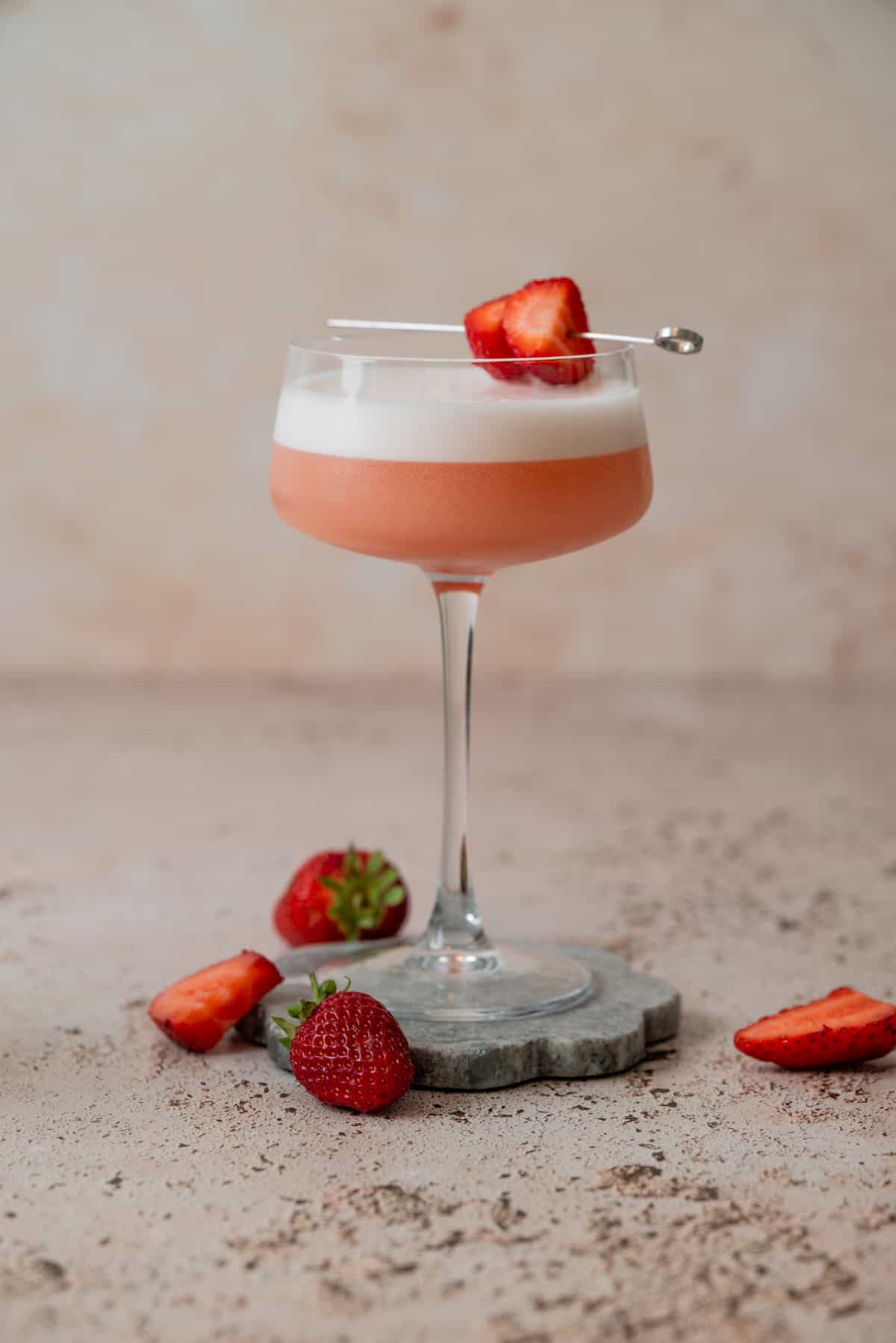 A pink Strawberry Gin Sour with a foamy top served in a coupe glass, garnished with sliced strawberries on a pick. Whole and sliced strawberries are scattered around the base on a light, textured surface.