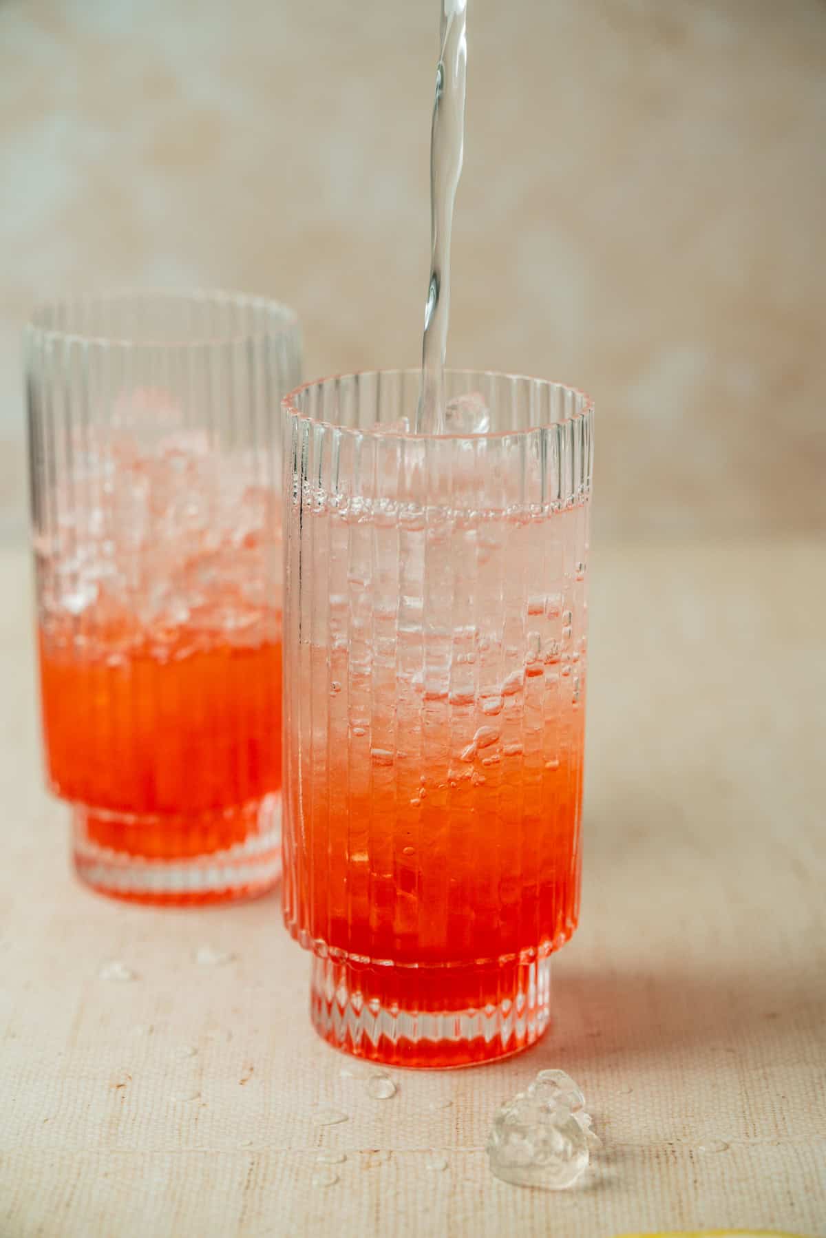 A clear liquid is being poured into a tall glass with vertical ridges, containing strawberry lemonade mocktail and ice. Another similar glass is in the background, with a heart-shaped ice cube on the surface nearby.