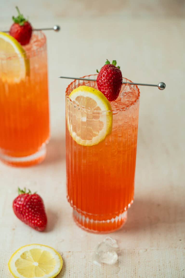 Two tall glasses filled with a bright orange-red strawberry lemonade mocktail, each garnished with a lemon slice and a whole strawberry on a skewer. Extra strawberries and lemon slices lay on the light surface nearby.