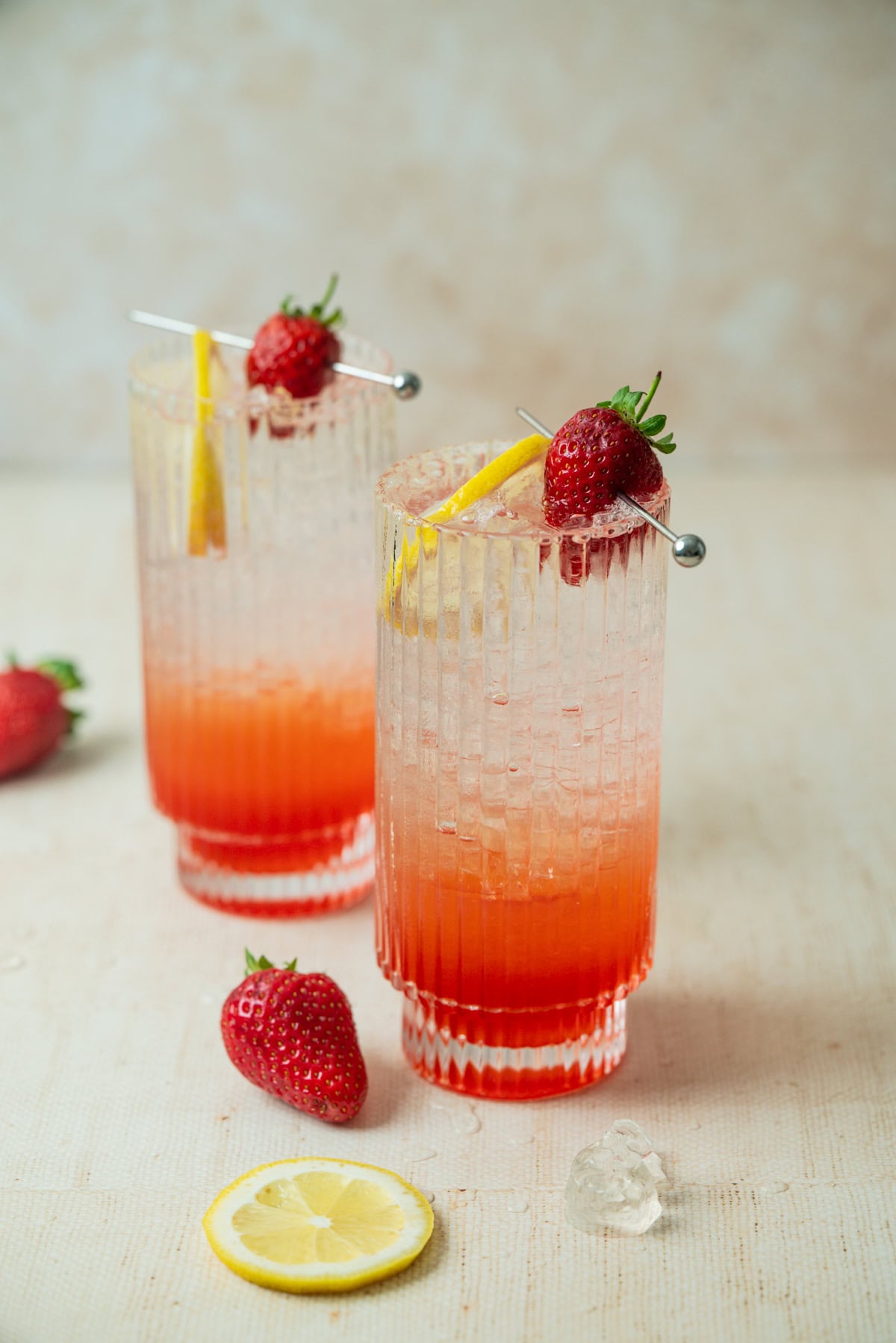 Two tall glasses filled with a red and clear layered strawberry lemonade mocktail, garnished with a strawberry and lemon slice. Extra strawberries, a lemon slice, and an ice cube rest on the light-colored surface nearby.