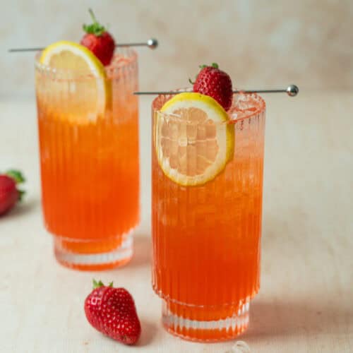 Two tall glasses of strawberry lemonade mocktail with ice, garnished with a lemon slice and a fresh strawberry. Two strawberries and a lemon slice rest on the light surface nearby. The neutral background is softly blurred.