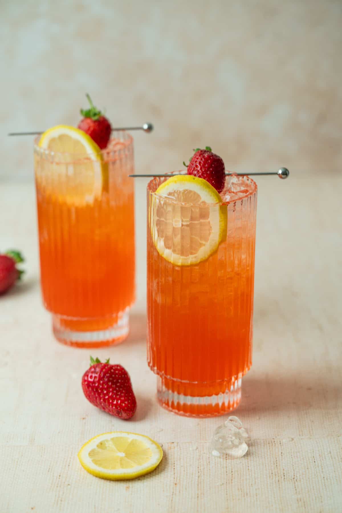 Two tall glasses of strawberry lemonade mocktail with ice, garnished with a lemon slice and a fresh strawberry. Two strawberries and a lemon slice rest on the light surface nearby. The neutral background is softly blurred.