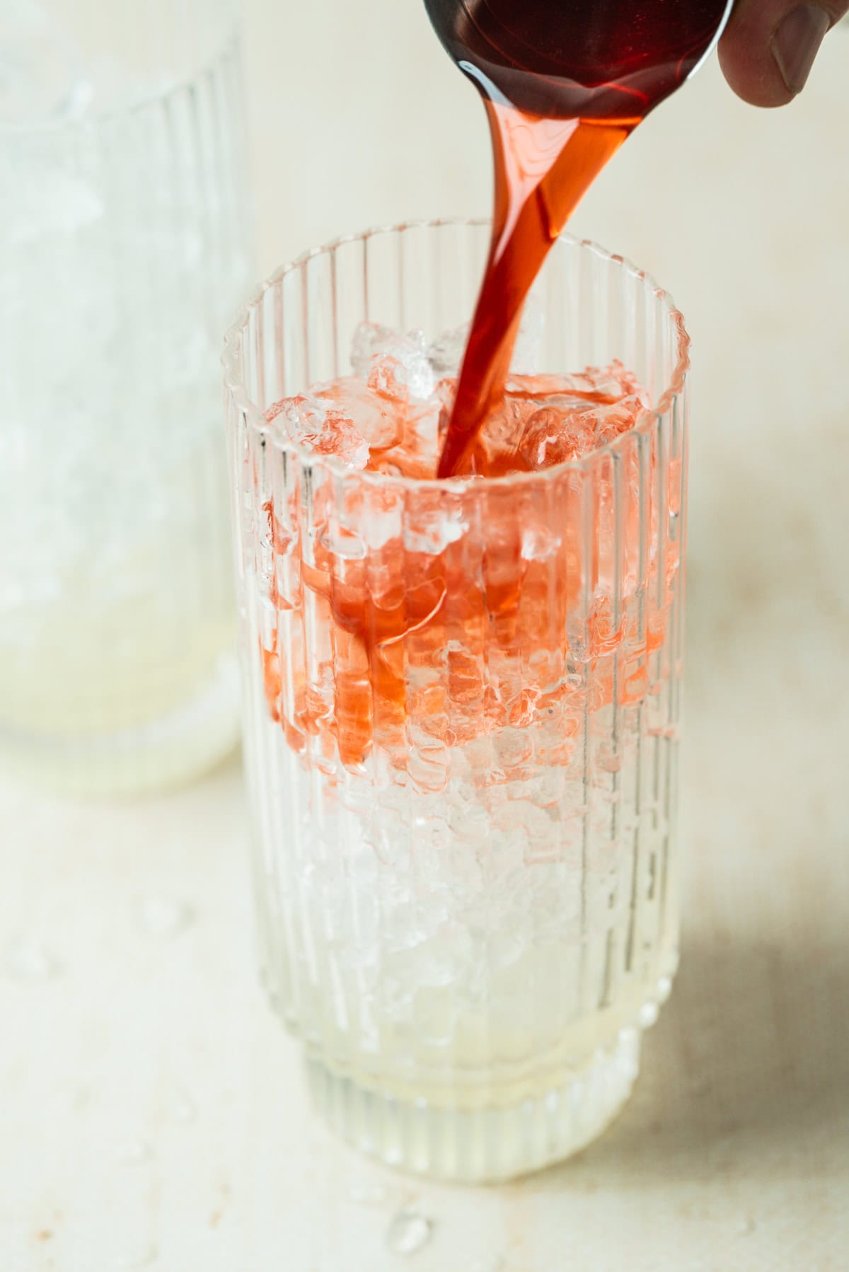 A close-up of a glass filled with crushed ice and pale liquid, as vibrant red strawberry lemonade mocktail is poured in, creating a swirling effect. Another glass is blurred in the background.