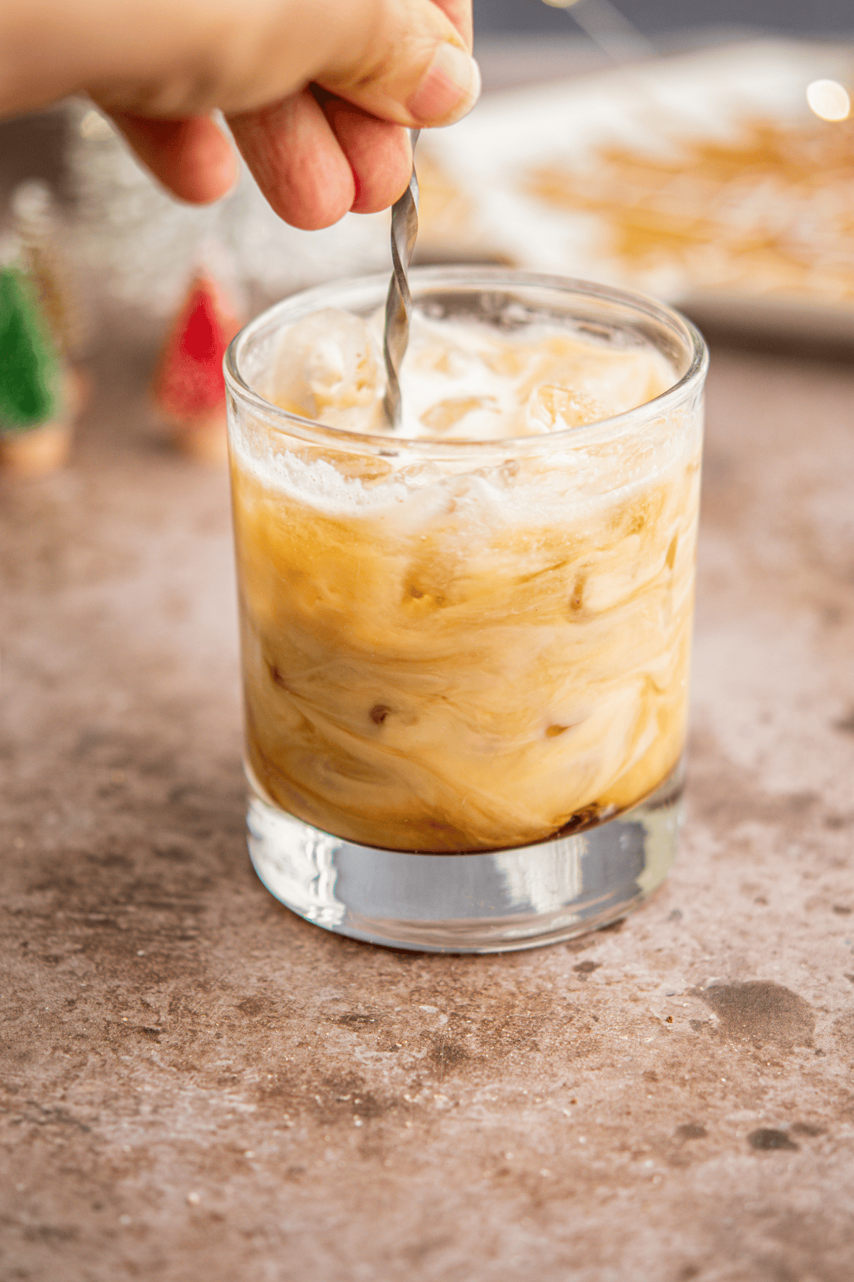 A hand stirs a creamy Eggnog White Russian in a clear glass, causing the milk and coffee to swirl together. The drink sits on a brown countertop with blurred holiday decorations in the background.