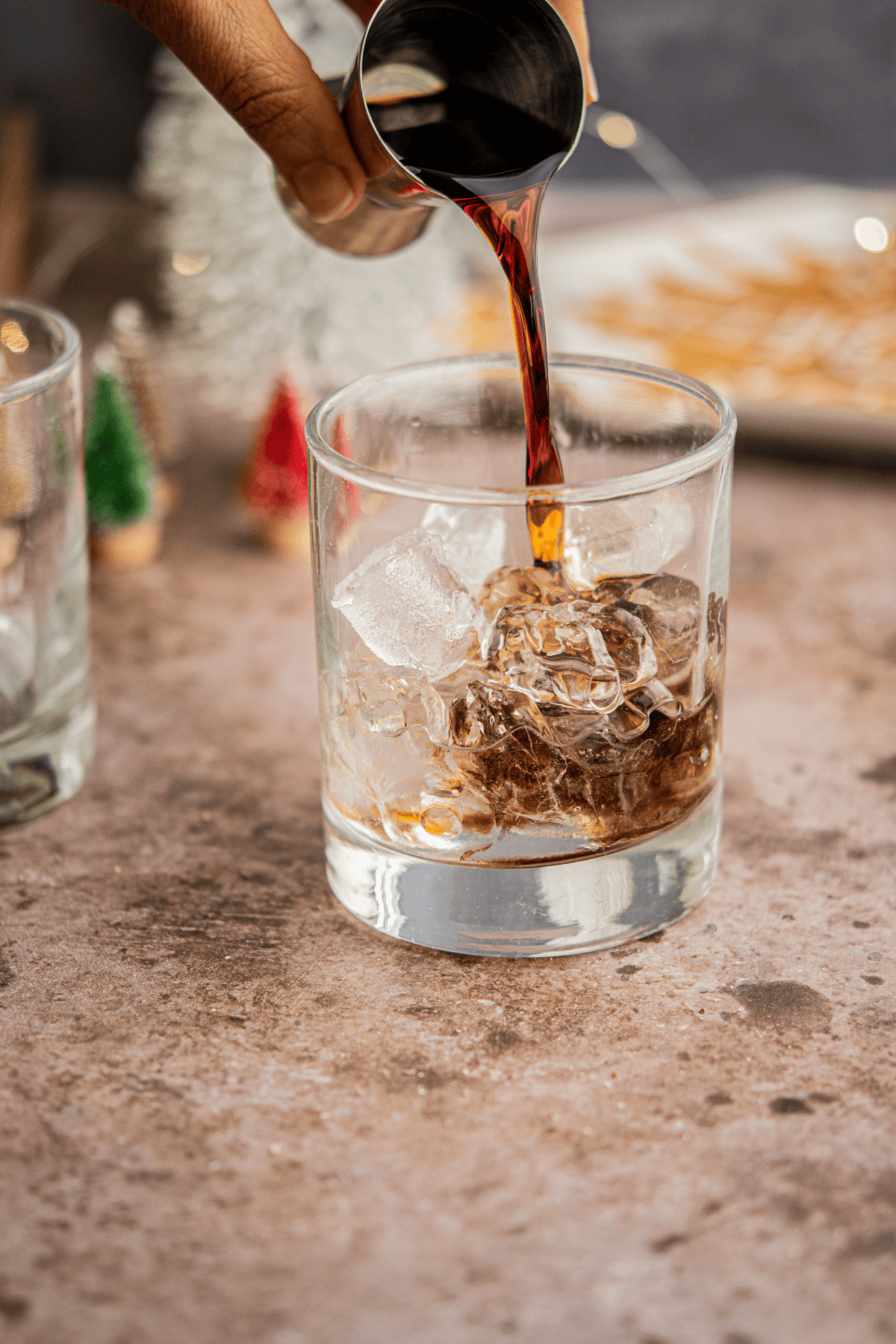 A hand pours a dark liquid from a metal jigger into a glass filled with ice cubes, crafting an Eggnog White Russian. Small holiday decorations and a plate of snacks are blurred in the background.