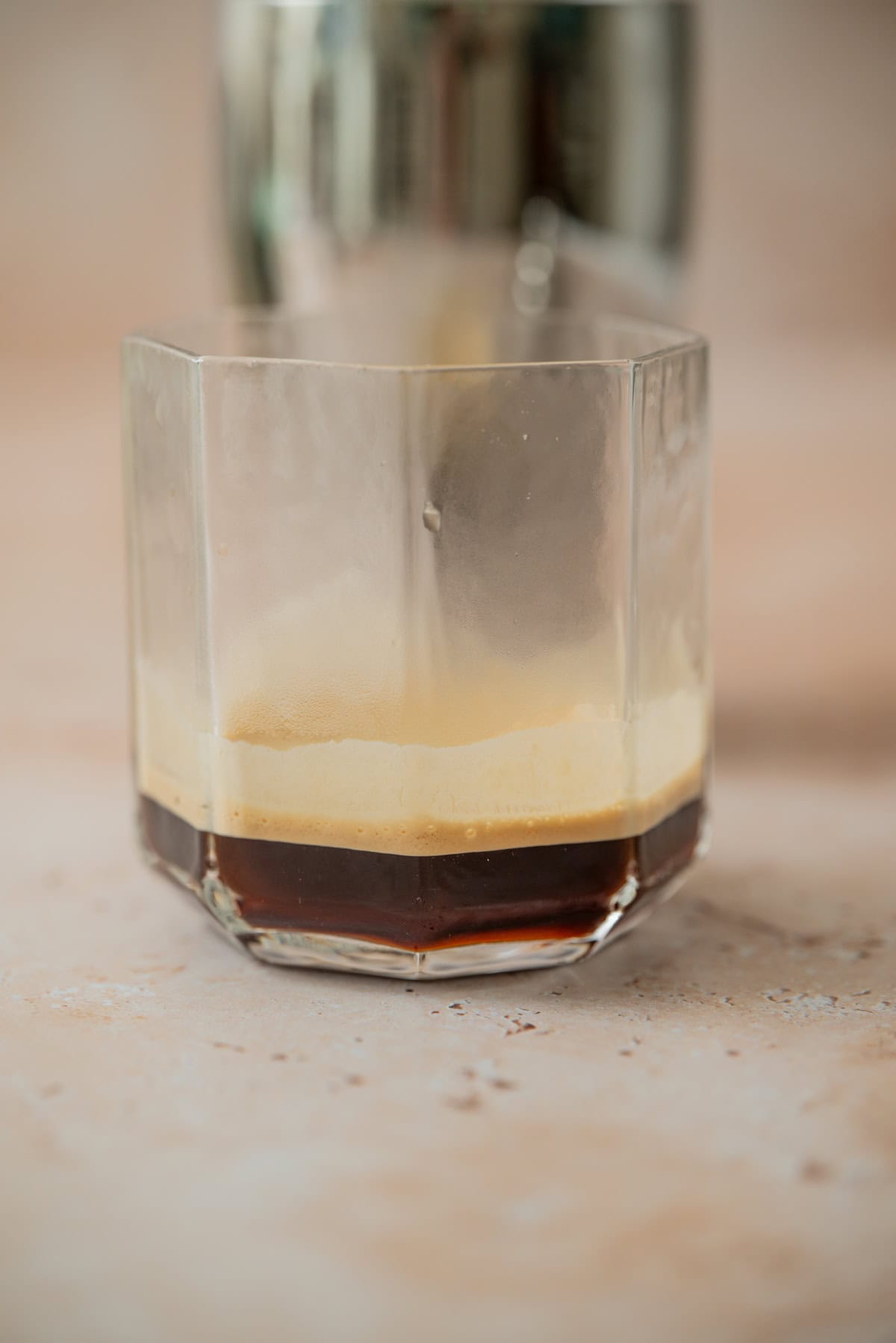 A close-up of a faceted glass with a dark coffee layer topped by light crema, reminiscent of an Eggnog Espresso Martini, sits on a beige surface with a blurred metallic object in the background.