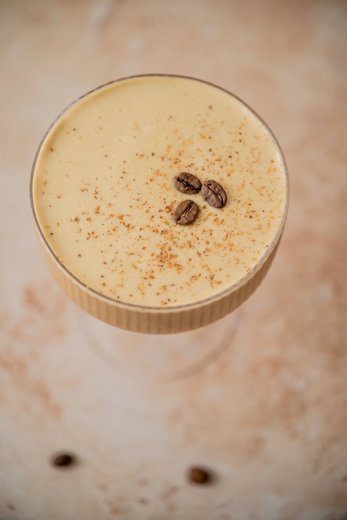 A creamy, coffee-colored Eggnog Espresso Martini in a glass, topped with three coffee beans and a light dusting of ground spice, viewed from above on a beige surface.