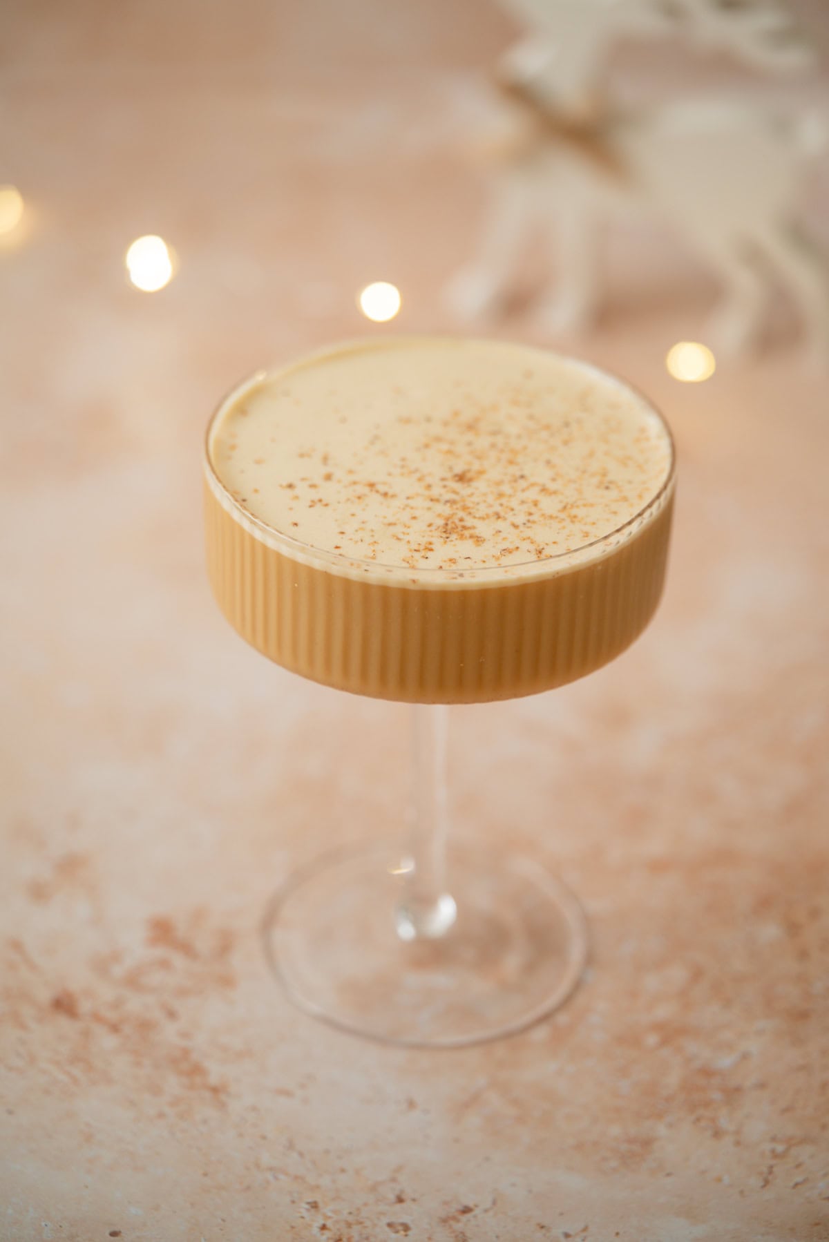 A creamy Eggnog Espresso Martini, topped with a sprinkle of nutmeg, served in a textured glass coupe on a light, speckled surface with blurred festive lights in the background.